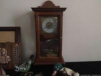 Photo showing vintage wooden wall clock with glass front and Roman numeral dial, with some ceramic duck figurines in front.