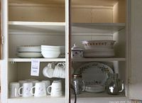 Entire cabinet view showing top shelves empty, middle shelves with stacked white plates and bowls, lower shelves with mugs and glassware