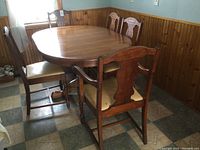 Photo of the full dining table with five chairs arranged around it in a room with wood paneling and patterned tile floor.