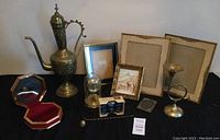 Full lot spread showing brass carafe, multiple picture frames, jewelry box, anniversary clock, brass trumpet candle holder, magnifying glass, and ladle.
