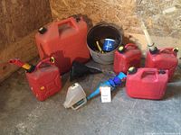 Six red gasoline cans of varying sizes with yellow/black caps on a concrete floor next to a gray bucket containing automotive items and several funnels.