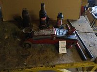 Showing entire lot with three hydraulic bottle jacks and one red hydraulic floor trolley jack on a wooden table, plus two metal handles.