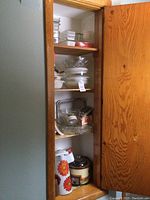 View of pantry shelves showing glass food storage containers, glass bowls, clear glass trays, and a vintage metal crockpot with flower decal graphic on lower shelf.