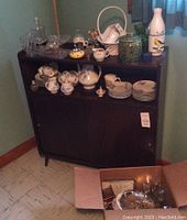 Small dark wood buffet with porcelain dinnerware and various glass and ceramic items on top and inside the cabinet.