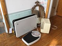 White space heater, Bonaire air purifier, ornate mirror, coffee table glass leaning against wall, and mechanical weight scale on wooden floor