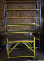 Front view of steel shelving unit and yellow workbench with casters inside a workshop with wooden walls.