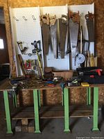 Full view of the green metal framed work bench with pegboard behind it. Several hand saws and other tools hanging on the pegboard and placed on the bench surface.