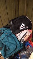Photo showing teal Protege framed hiking backpack, black Adidas duffel bag, small pink and gray bag and other bags stacked together.