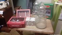 Photo shows clear glass tree-shaped candy jars, boxed kitchen utensil ornaments with visible spoons and forks, and boxed holiday ornaments on a wooden table.