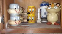 Photo of collectible character cups including two Bugs Bunny, two Tweety Bird, two Wile E. Coyote, one Meowth, and one R2D2 cup on wooden shelf with visible dust and wear.