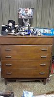 Front view of 5 drawer wooden dresser showing scratches and wear on surface, metal handles on drawers, and tapered mid-century style legs.