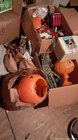 Multiple cardboard boxes and paper bags with orange plastic pumpkin buckets, black light boxes, string lights, flying bats decorations, and an animated shrieking ghost box.