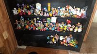 Three shelves filled with many small vintage toy figurines from McDonald's and Burger King kids meals along with Barbie dolls. Various cartoon and movie characters are visible with varied colors and sizes.