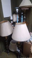 Photo showing several lamps with beige lampshades, including tripod wooden lamp and darker wood base lamps with shades present
