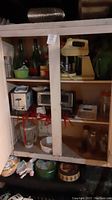 Cabinet showing vintage hand mixer, toaster, toaster oven, large green bottles, glass jars, pitcher, and planter.