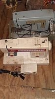 Photo showing two sewing machines placed on wooden surface. One is Simplicity branded with red and beige accents, the other is a gray Sears machine. Both are dusty and untested.