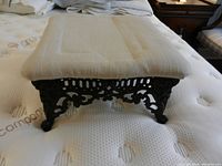 Close-up of foot stool showing ornate black iron base with intricate scrollwork and beige upholstered cushion top.