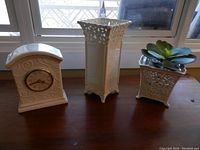 Ivory ceramic clock, tall and small filigree vases displayed on wooden surface near window, highlighting filigree details and clock face