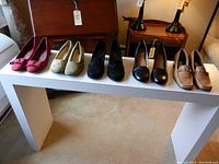 Photo shows five pairs of women's shoes in different styles and colors arranged on a white bench. Colors include pink, beige, black, and tan. Styles include flats and heels.