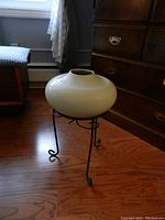 View of the ceramic vase placed on black metal stand on wood floor, next to dresser and chair