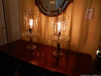 Pair of lit cherub style lamps with cut glass bases and hanging glass prisms on a wooden surface.