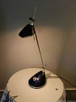 Blue and silver desk lamp shown lit on a table against a plain wall. The lamp emits warm light downward with a conical shade.