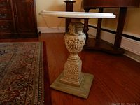 Full view of the pedestal table showing the white round marble top and detailed carved base on a wooden floor beside other furniture.