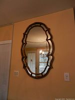 Front view of ornate gold painted wood frame wall mirror hanging on beige wall, showing the full oval mirror shape and scalloped edges.