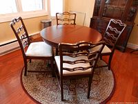 Full view of round pedestal dining table with 4 ladderback chairs with cream upholstered seats, sitting on a round patterned rug.