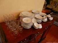 Photo showing water glasses and stacks of plates, bowls, cups, and serving pieces arranged on wooden surface