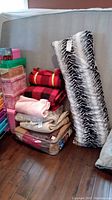 Full view of multiple blankets, throws, a pillow and a comforter stacked near a wall on wooden floor. Includes zebra print, red and black plaid and other solid color fabrics.