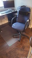 Blue adjustable office chair placed on clear plastic floor protector in home office setting with desk, keyboard, printer, and monitor in background.