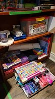 Photo showing shelves with crafting supplies including plastic storage bins, paint palettes, and various boxes of Rainbow Loom kits and elastics on floor and shelf.