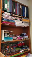 Four wooden shelves filled with assorted crafting and stationery supplies including binders, loose paper, pencil cases, art materials, and calculators