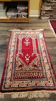 Full view of hand knotted rug placed on wood floor, showing geometric and floral design with red center and cream border.