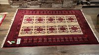 Full view of rectangular hand knotted Persian rug with red border and cream center field, laid out on floor.