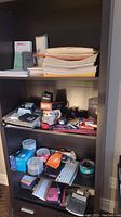 Photo showing shelves with stacks of paper, envelopes, folders, boxes, electronic items, two keyboards, disc spindles, and office supplies scattered on shelves.