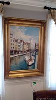 Full view of framed oil painting hanging on wall near window, showing canal scene with boats and buildings under cloudy sky.