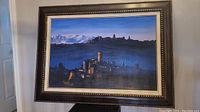 Framed painting showing the scenic town with mountain background and clear sky.