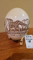 Front view of ostrich egg showing giraffe and tree hand painted details on the egg mounted on a three-legged stand
