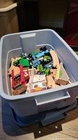 Gray plastic storage bin containing multiple Thomas and Friends wooden trains, tracks and accessories, shot from a top angle.