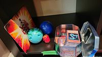 Yu-Gi-Oh cards stored in a Pokemon Diamond & Pearl metal card tin alongside three Bakugan figures (green, blue and small red) and a Bakugan arena/fold-out mat.