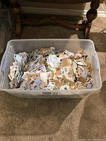 Large plastic storage tub filled with numerous international stamps on paper pieces, shown from the front with varied colorful stamps visible.