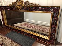 Full view of the horizontal rectangular mirror showing the dark reddish-brown carved floral frame and gold inner border laid on the floor.