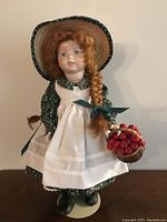 Front view of Anne of Green Gables porcelain doll wearing green floral dress, white apron, straw hat, holding a basket of cherries.