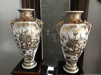 Pair of tall ivory colored Satsuma pottery vases with detailed floral and gilt decorations, displayed on black stands under warm lighting
