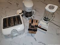 Photo showing T-Fal toaster, Westinghouse coffee maker, Osterizer blender, and Starfrit knife block with knives arranged on a tile floor.