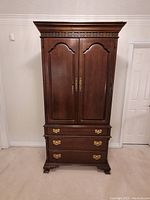 Front view of large hardwood armoire showing two carved doors, brass handles, and three drawers below.