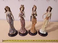Front view of four female figurines in detailed long dresses with floral patterns, standing side by side on round bases, measuring tape visible for scale.