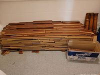 Stack of used hardwood flooring planks with several solid wood spindles and posts leaning against the wall on the right. A cardboard box is placed next to the stack.